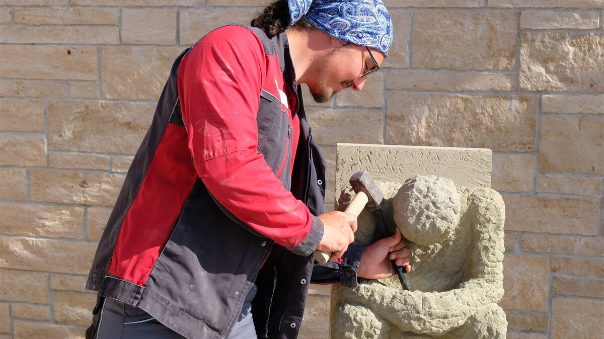 Leonhard arbeitet mit Hammer und Meißel an einer Steinskulptur in Form eines Menschen.