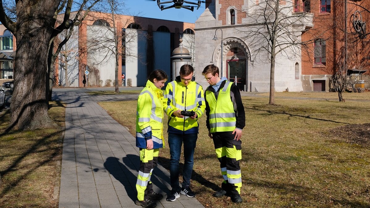 Doro, Peter und ihr Ausbilder in Warnkleidung im Freien mit Drohnensteuergerät und Drohne.