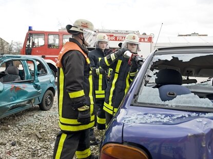 Drei Feuerwehrkräfte zerschlagen die Scheiben eines Autos.