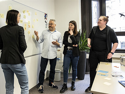 Vier Personen stehen vor einem Whiteboard und besprechen etwas.