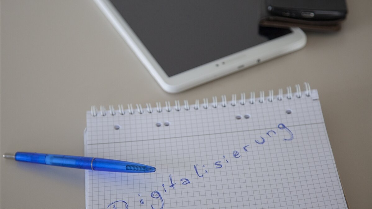 Ein Block, auf dem das Wort "Digitalisierung" steht, liegt neben einem Tablet und einem Handy auf einem Tisch.