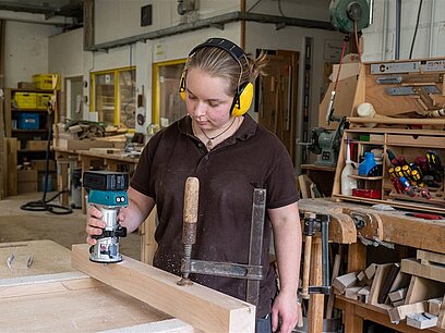 Eine junge Frau mit Gehörschutz bearbeitet ein Holzteil mit einer Hobelmaschine.