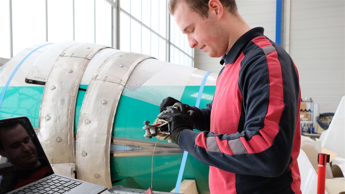 Ein junger Mann mit Schutzkleidung und Handschuhen testet ein Flugzeugbauteil auf Funktionalität.