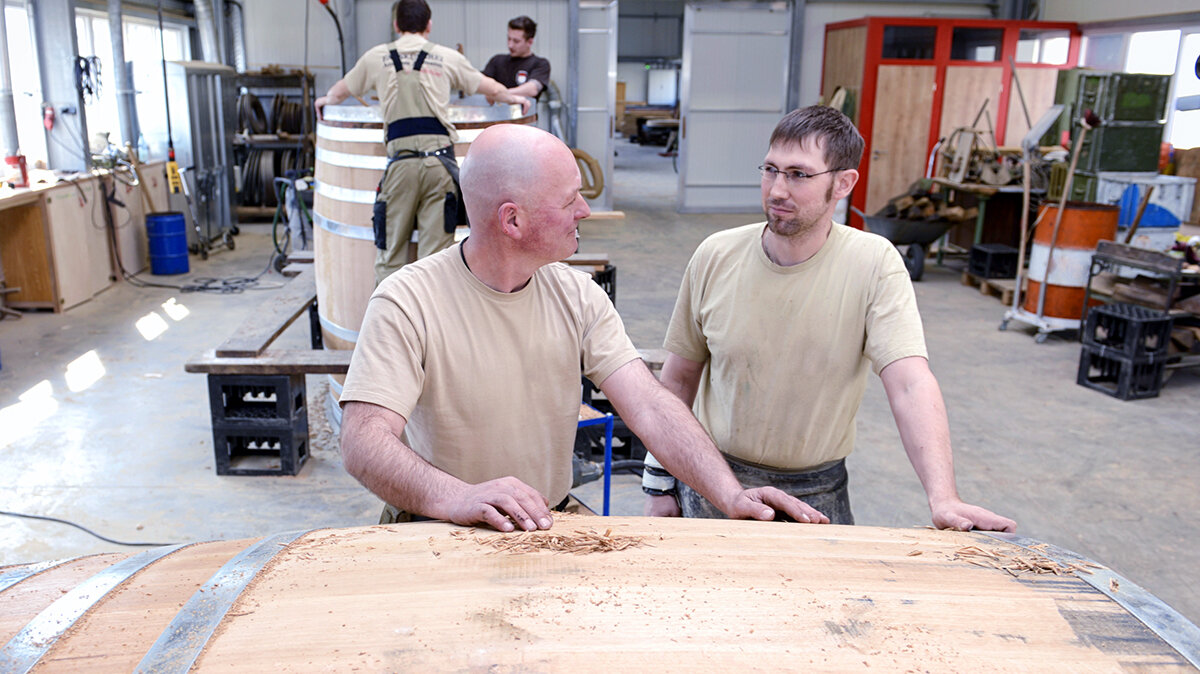 Zwei Männer stehen in einer Arbeitshalle vor einem Holzfass und unterhalten sich.