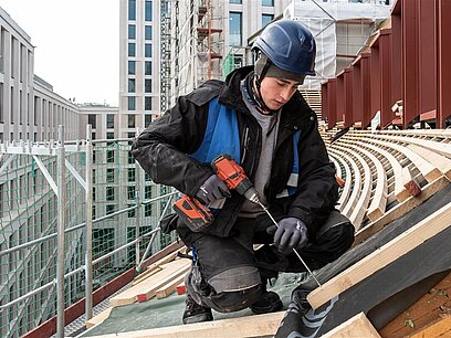 Ein junger Mann mit Helm und Arbeitskleidung schraubt Holzbalken auf einem Dach fest.