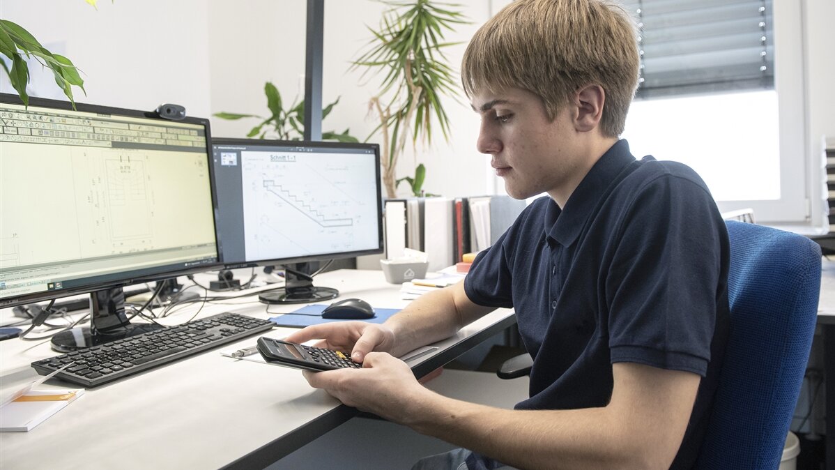 Ein junger Mann sitzt an seinem Arbeitsplatz und rechnet mit einem kleinen Taschenrechner.