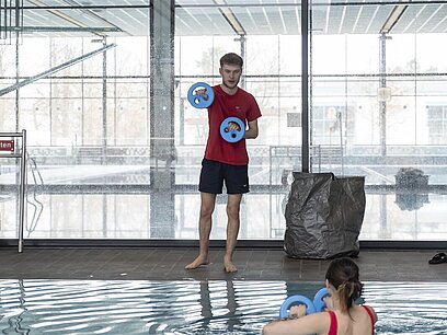 Ein junger Mann mit Wasserhanteln in den Händen steht vor einem Schwimmbecken und trainiert eine junge Frau im Wasser mit Wasserhanteln in den Händen.