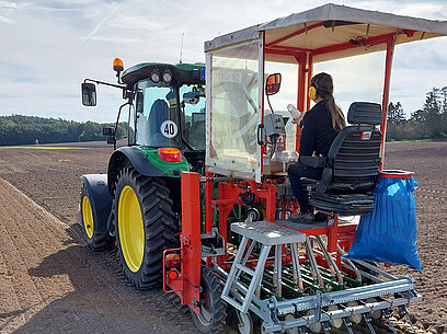 Eine Frau mit Gehörschutz sitzt auf dem Anhänger eines Traktors auf einem Feld.