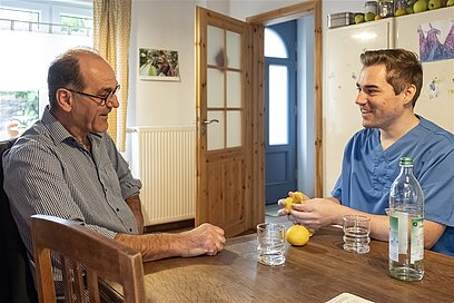 Ein junger Mann in Pflegekleidung sitzt mit einem älteren Herrn am Tisch und schält eine Orange.