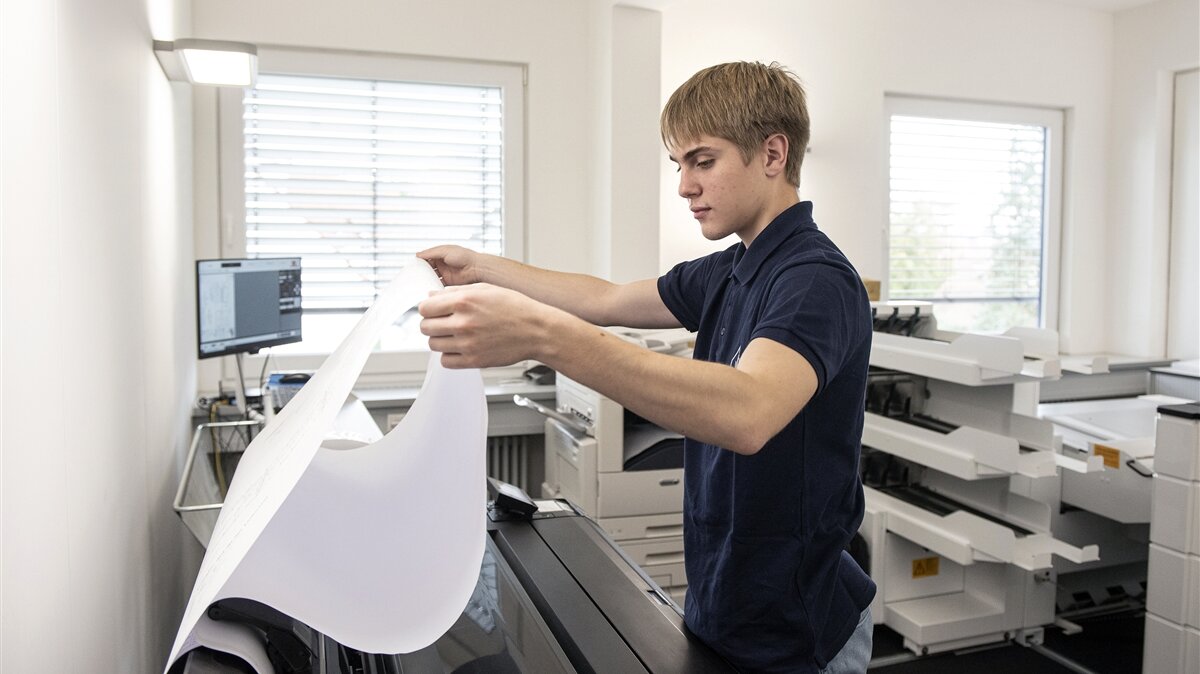 Ein junger Mann nimmt mit beiden Händen eine große Zeichnung aus einem großen Drucker.