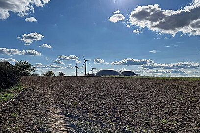 Hinter einem Acker stehen Windräder und eine Biogasanlage.