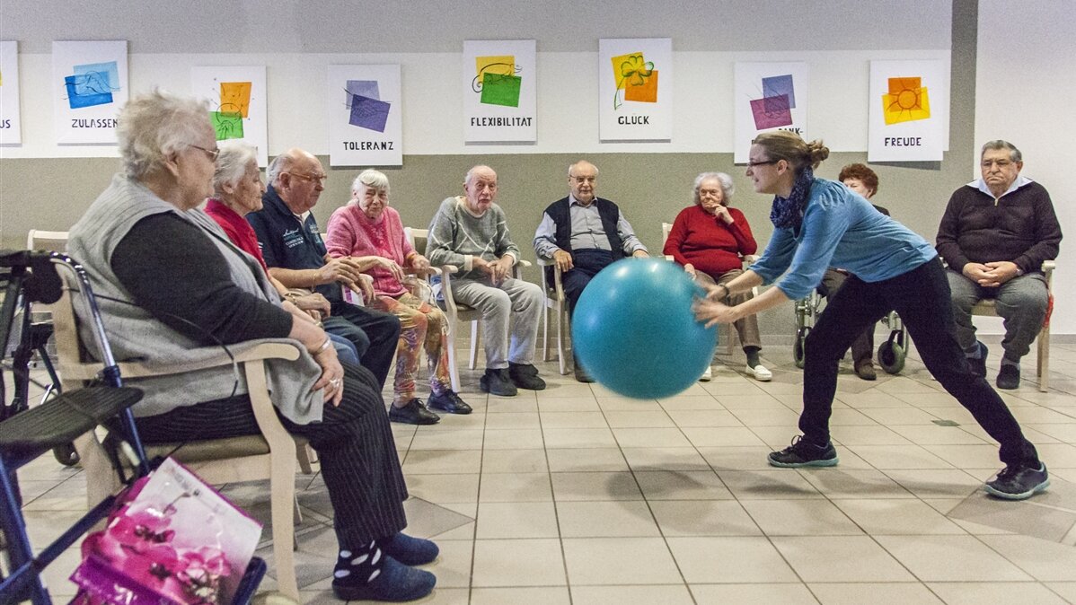 Eine junge Frau aktiviert Senioren in einem Pflegeheim mit einem Ballspiel. 