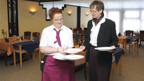Eine Ausbilderin erklärt einer Auszubildenden im Gastgewerbe, wie sie die Teller hält zum Servieren.