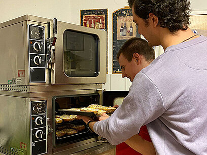 Mate unterstützt einen Bewohner dabei, ein Blech in den Ofen zu schieben.