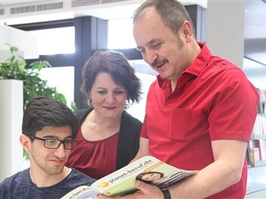 Ein Vater, eine Mutter und ein Sohn lesen zusammen in einem Heft von planet-beruf.de.