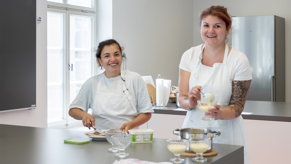 Zwei junge Frauen in Schürzen bereiten Essen in einer Küche zu.