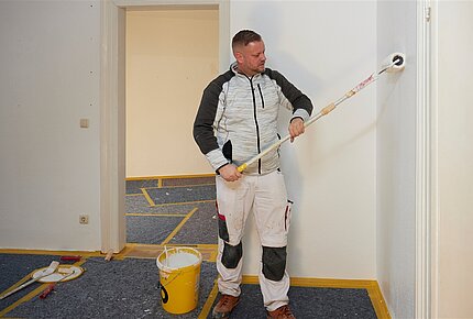 Ein junger Mann in Arbeitskleidung streicht mit einer Farbrolle eine Wand. 