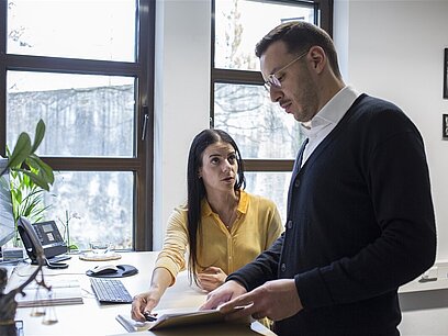 Eine Frau und ein Mann deuten auf etwas in einer Akte am Schreibtisch in einem Büro.