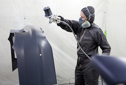 Eine junge Frau in Schutzkleidung und Mundschutz lackiert mit einer Farbpistole ein Autoteil. 
