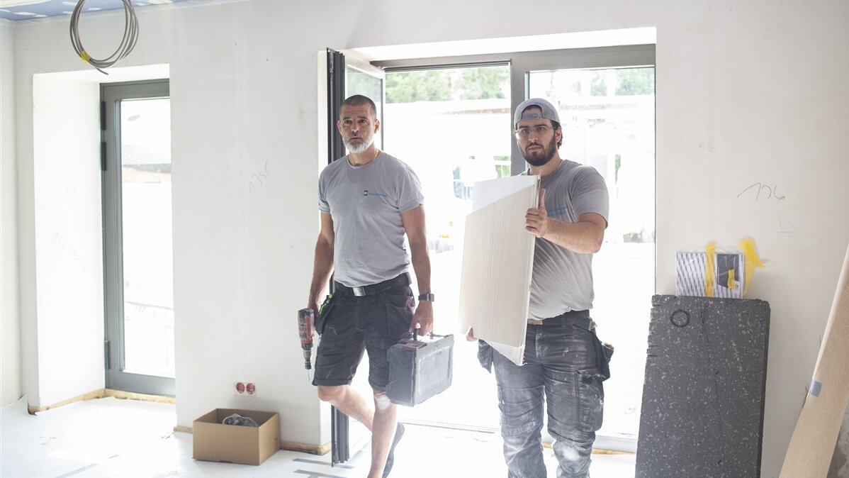 Niklas trägt mit einem Kollegen eine Gipskartonplatte und ein Bohrmaschine ins Haus.