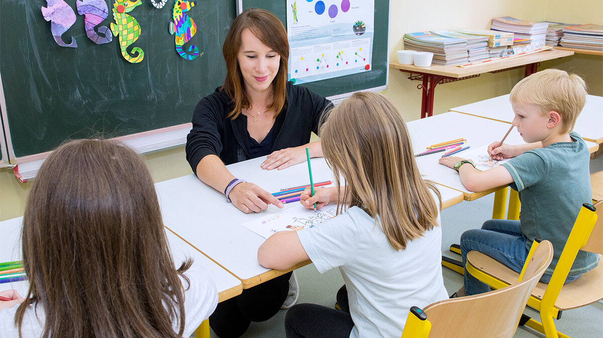 Eine junge Frau sitzt mit Kindern, die malen, an einem Tisch.