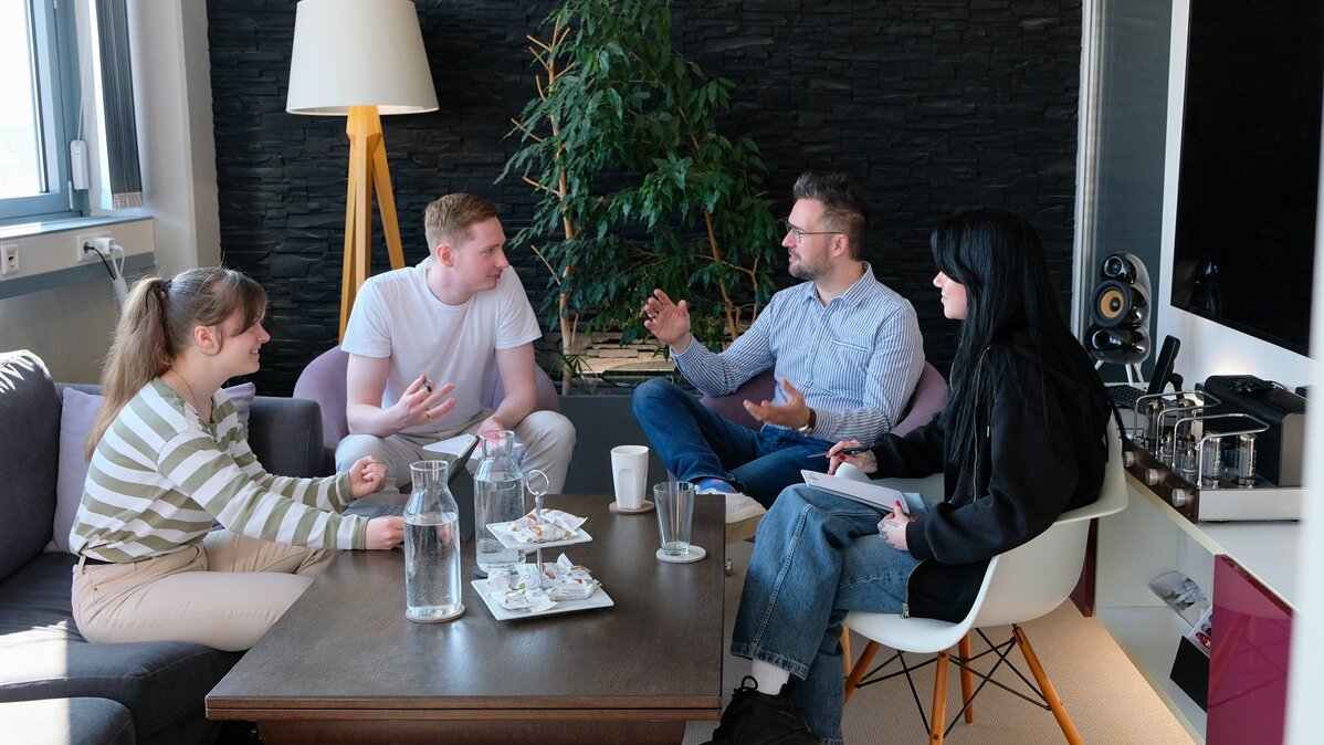 Ein junger Mediengestalter spricht mit Kolleginnen und Kollegen im Besprechungsraum.
