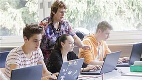 Eine Lehrerin erklärt drei Jugendlichen etwas, die in einer Reihe vor ihren Laptops sitzen.