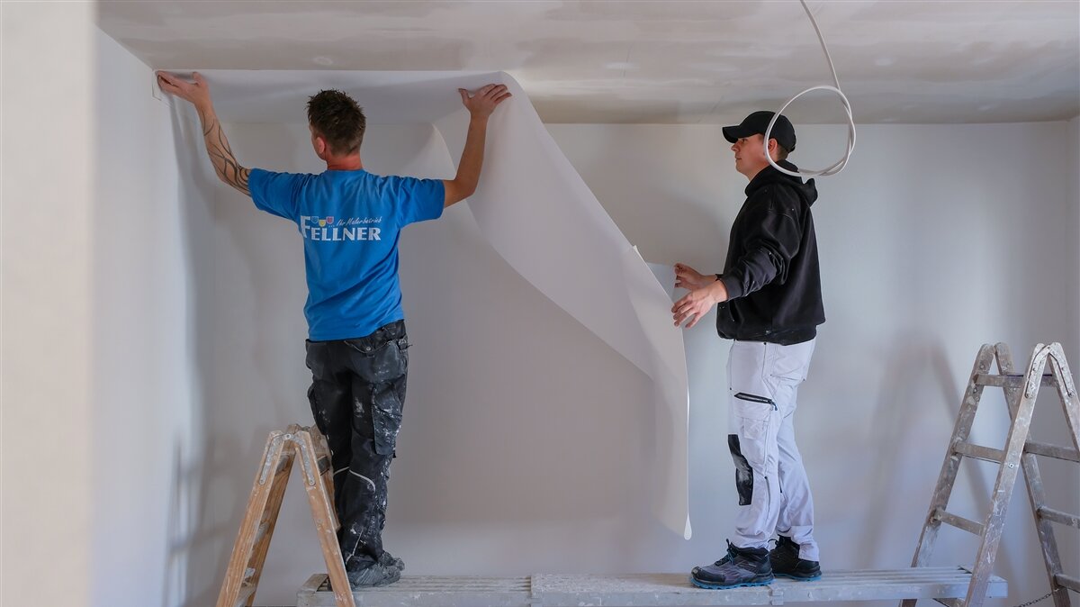 JJulian und sein Kollege befestigen in Arbeitskleidung eine Tapetenbahn an einer Decke.