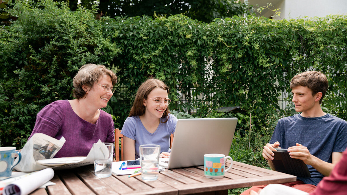 Eine Familie sitzt zusammen am Gartentisch und sie schauen in einen Laptop. 