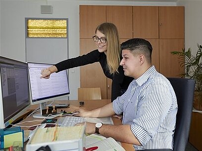 Ein junger Mann arbeitet am Computer, während ihm eine junge Frau etwas erklärt.