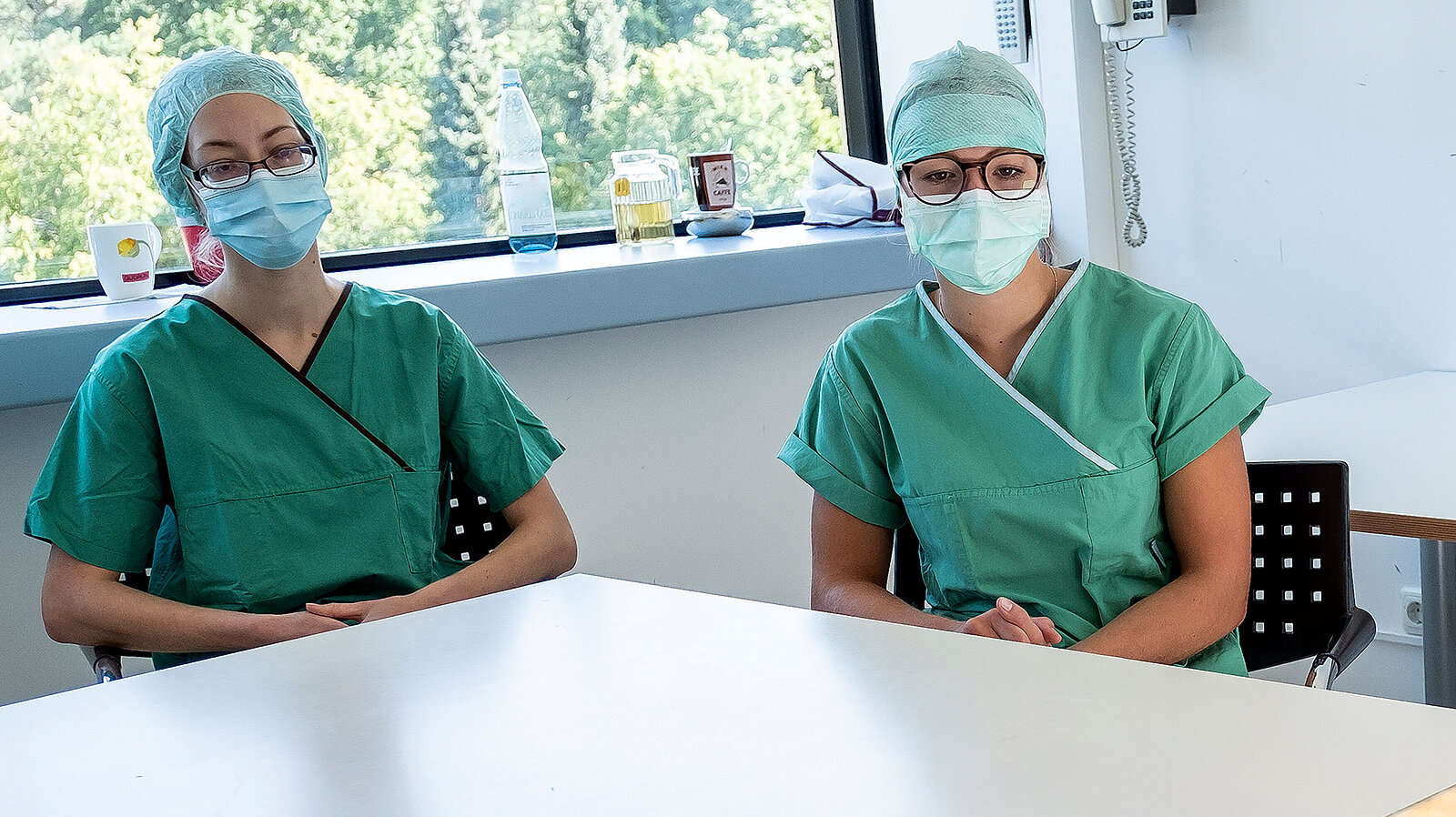 Zwei Frauen sitzen in OP-KLeidung an einem Tisch und schauen in die Kamera.
