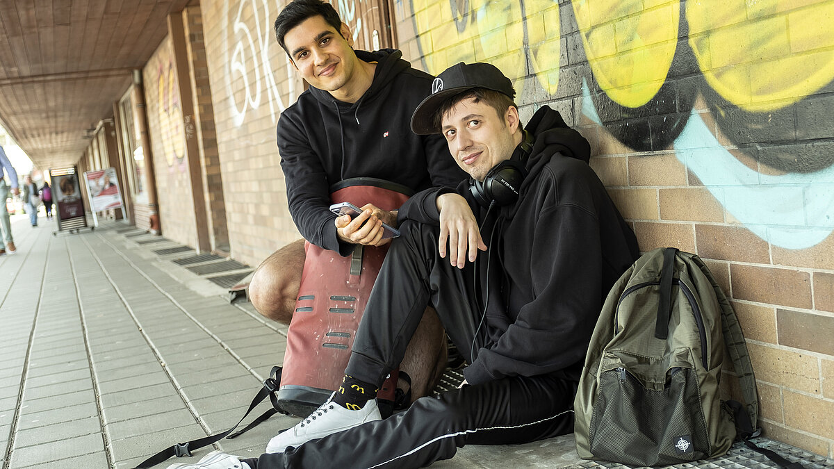 Zwei junge Männer sitzen mit Rucksäcken und Handy vor einer Wand und schauen in die Kamera.