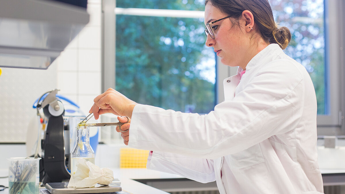 Ein Frau im Laborkittel schneidet eine Probe in ein mit Flüssigkeit gefülltes Glas.