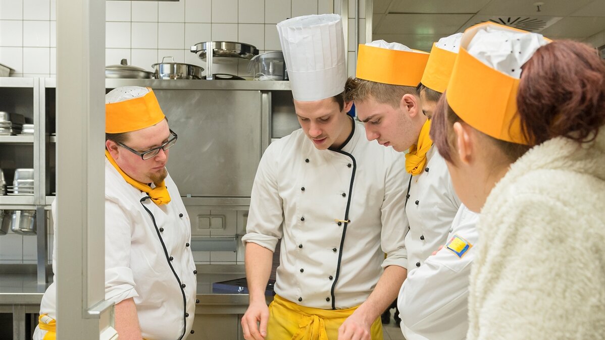 Ein Koch unterweist vier Auszubildende in einer Gastronomieküche.