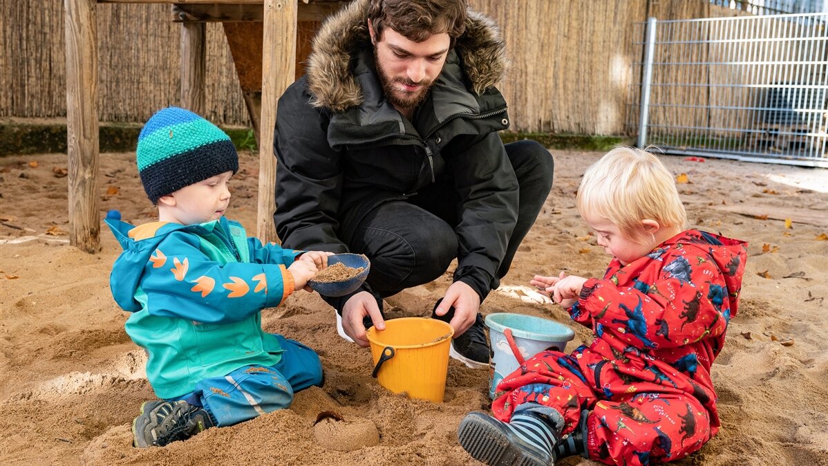 Ein junger Mann spielt mit zwei Kindern im Sandkasten.