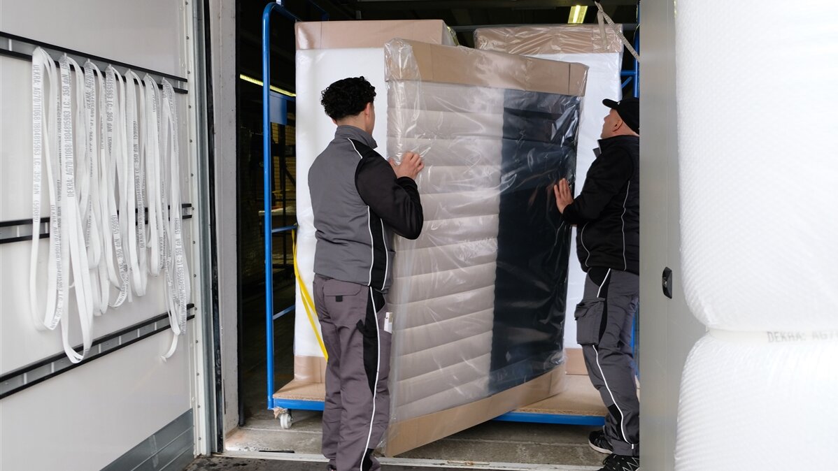 Zwei junge Männer schieben ein großes verpacktes Möbelstück in einen Rollbehälter.