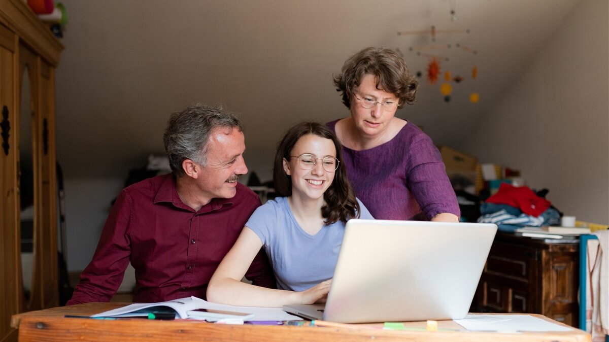 Eine Mutter und ein Vater unterstützen ihre Tochter beim Arbeiten am Laptop.