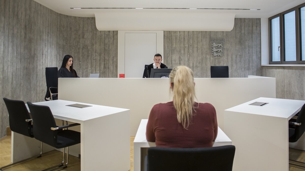 Ein Mann und eine Frau in Robe sitzen hinter einem Pult und eine Frau sitzt davor an einem Tisch im Gerichtssaal.