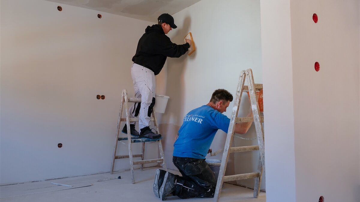 Julian, auf einer Leiter, und sein Kollege bearbeiten mit Kellen und Putz eine Wand eines Wohnraums.