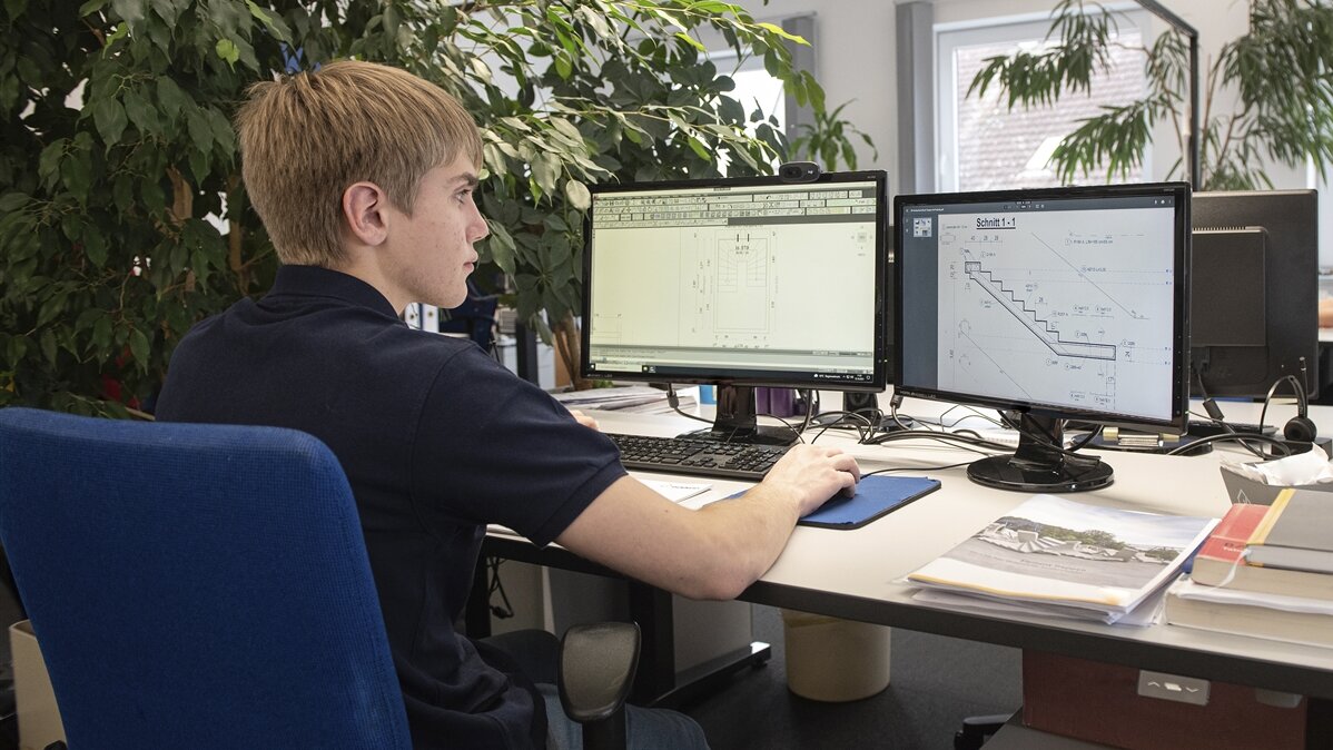 Ein junger Mann sitzt an seinem Arbeitsplatz und blickt auf zwei Bildschirme.