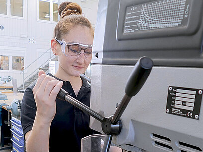 Anna-Maria trägt eine Schutzbrille und arbeitet an einer Maschine mit einem Drehkreuz.