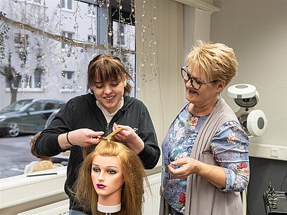 Eine junge Frau schneidet unter Anleitung die Haare einer Übungspuppe.