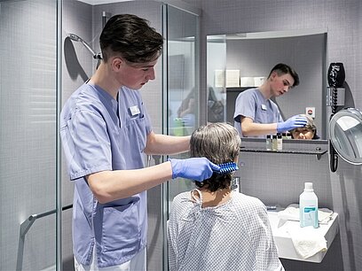 Ein junger Mann in einem Pflegekittel hilft einer Frau und bürstet ihr Haar in einem Badezimmer.