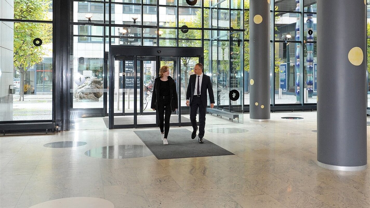 Ein Mann und eine Frau betreten ein Gebäude mit hoher Glasfront und Säulen