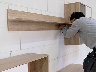 Ein junger Mann schraubt ein Brett an einer Wand fest.