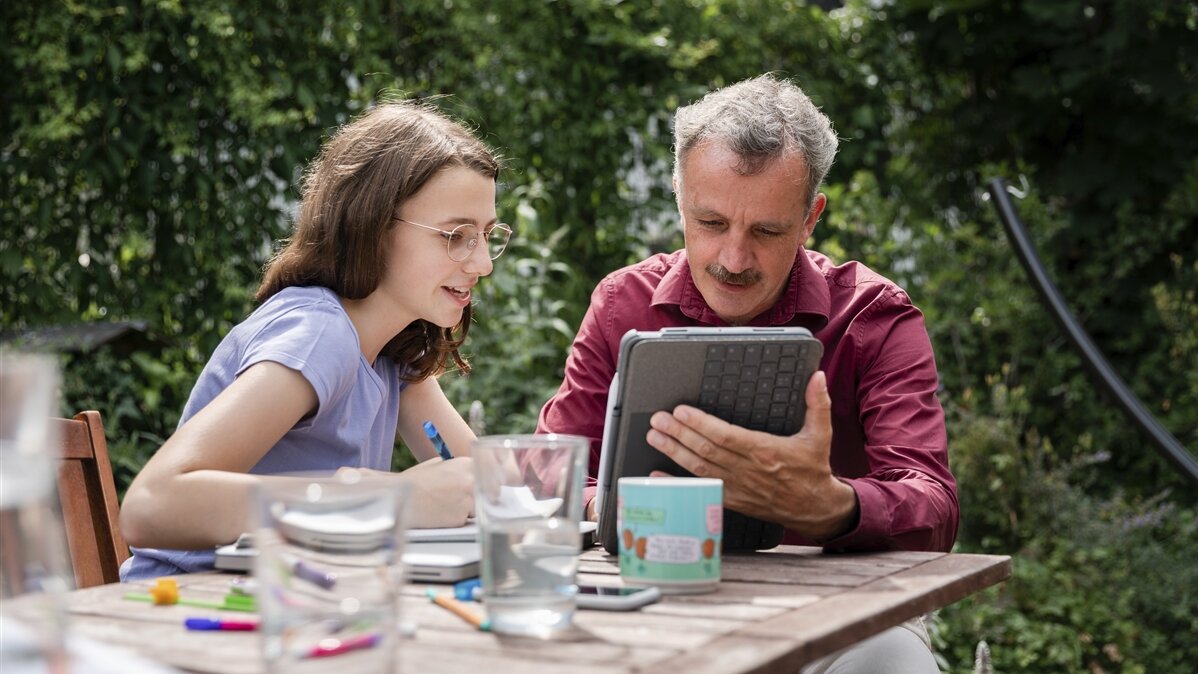 Ein Mädchen und ein Mann sitzen mit einem Tablet am Tisch im Grünen.