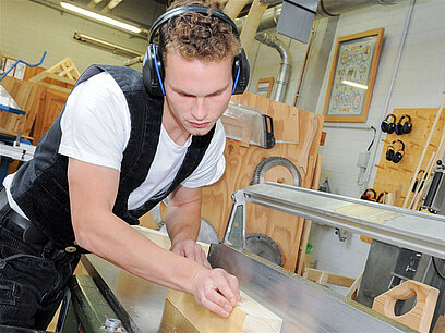 Ein junger Mann in Zimmererkleidung bearbeitet ein Holzstück an einer Maschine.
