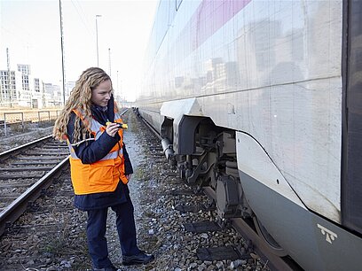 Eine junge Frau mit einer Weste prüft mit einer Taschenlampe einen Zug auf den Gleisen.
