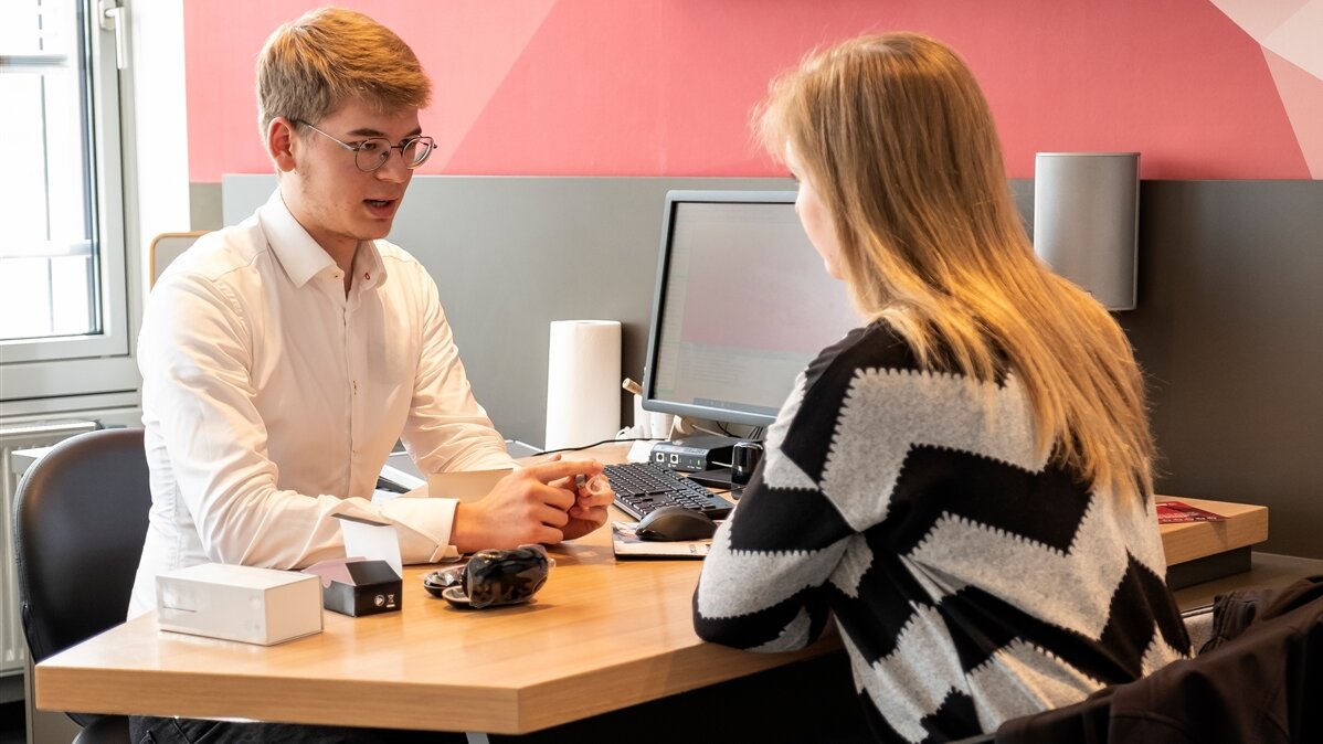 Ein junger Mann sitzt an einem Schreibtisch einer jungen Frau gegenüber und spricht mit ihr.