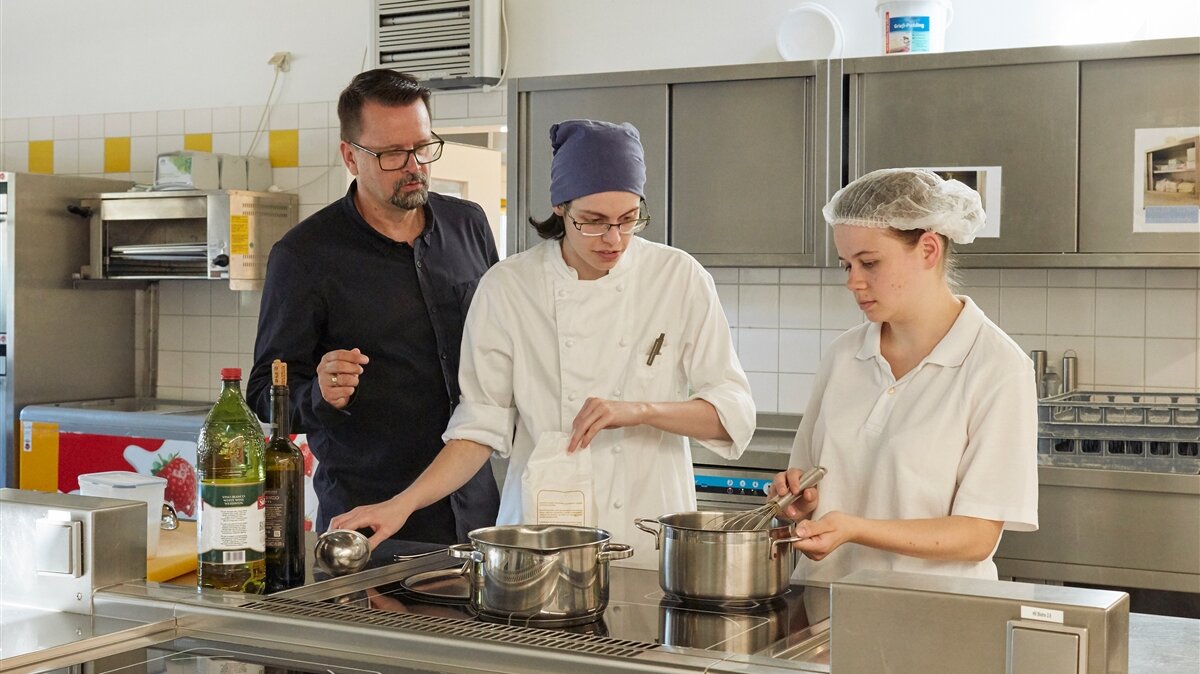 Zwei Männer und eine Frau stehen in einer Großraumküche und bereiten Essen in einem Topf zu.